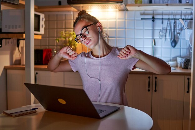 Mulher sonolenta, cansada e workaholic, usando óculos e fones de ouvido, alongando os músculos e fazendo uma pausa durante o trabalho árduo em um laptop à noite