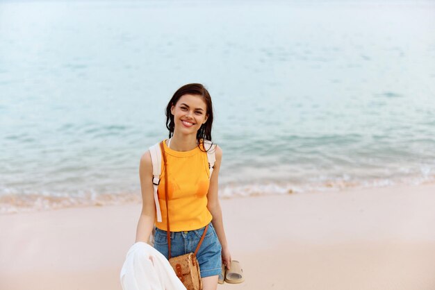 Mulher sorri com os dentes depois de nadar no oceano com uma mochila em um top amarelo molhado e shorts jeans caminha ao longo das férias de verão da praia em uma ilha à beira-mar no pôr do sol de Bali