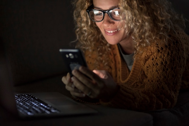 Mulher sorri usando conexão telefônica à noite no escuro em casa - pessoas e vício em dispositivos e tecnologia da web - uma bela mulher adulta procura e se comunica com o celular