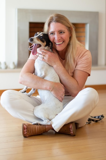 Mulher sorridente, abraçando o cão brincalhão. Mulher adulta mediana sentada no chão, segurando o cachorrinho branco. Amor, conceito de animal de estimação