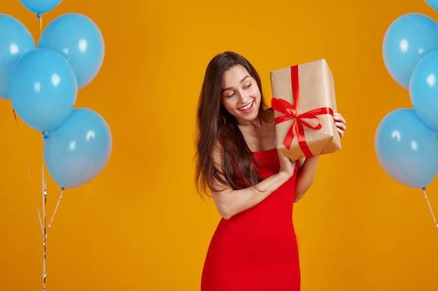 Mulher sorridente abre a caixa de presente com fitas vermelhas, fundo amarelo. Uma pessoa bonita do sexo feminino tem uma surpresa, evento ou festa de aniversário, decoração de balões