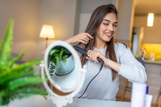 Mulher sorridente, alisando o cabelo com alisador de cabelo em casa.