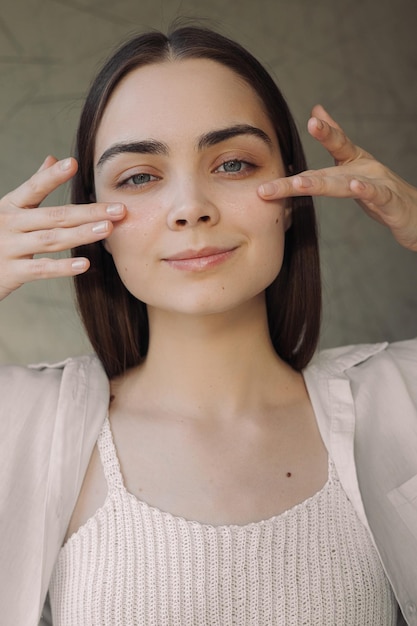 Mulher sorridente aplicando creme para os olhos e fazendo rotina de beleza em casa enquanto olha para a câmera