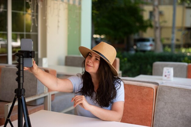 Mulher sorridente atraente do lado de fora sentado com tripé e smartphone