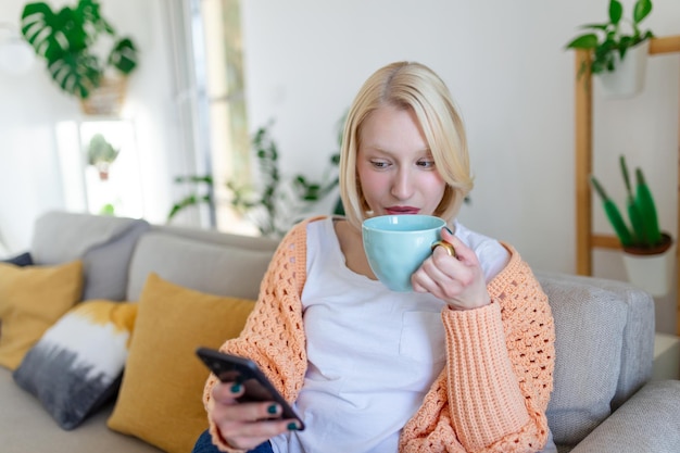 Mulher sorridente atraente usando telefone inteligente enquanto está sentado no sofá em casa conceito de comunicação e aconchego