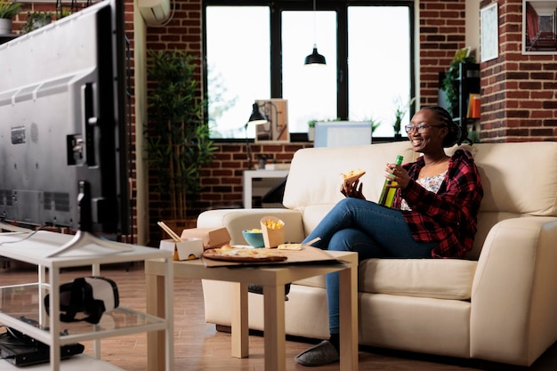 Mulher sorridente bebendo cerveja de garrafa e comendo pizza, assistindo filme no programa de televisão com refeição de entrega para viagem. desfrutando de filme na tv e fast food com bebida alcoólica.