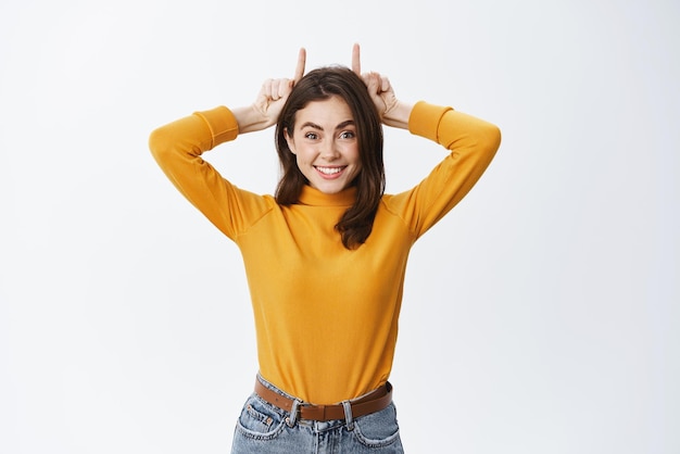 Mulher sorridente bonita mostrando chifres de dedo na cabeça e parecendo alegre se divertindo em pé brincalhão contra fundo branco