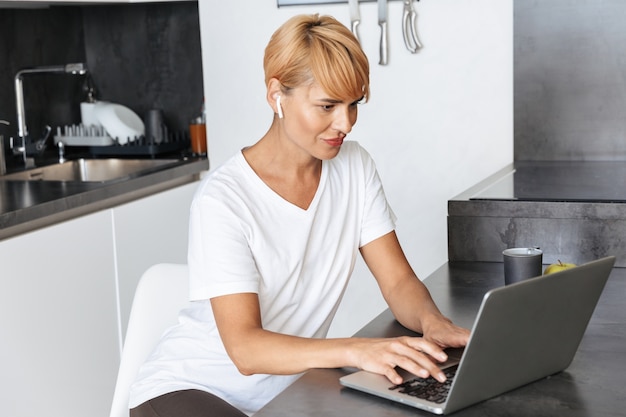 Mulher sorridente casual vestida usando um laptop enquanto está sentada na cozinha, usando fones de ouvido