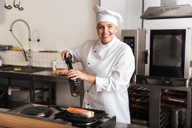 Mulher sorridente, chef cozinheira de uniforme, cozinhando um delicioso bife de salmão em pé na cozinha