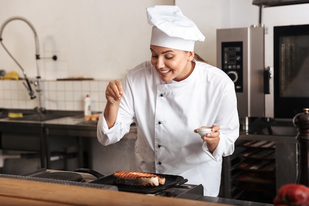 Mulher sorridente, chef cozinheira de uniforme, cozinhando um delicioso bife de salmão em pé na cozinha