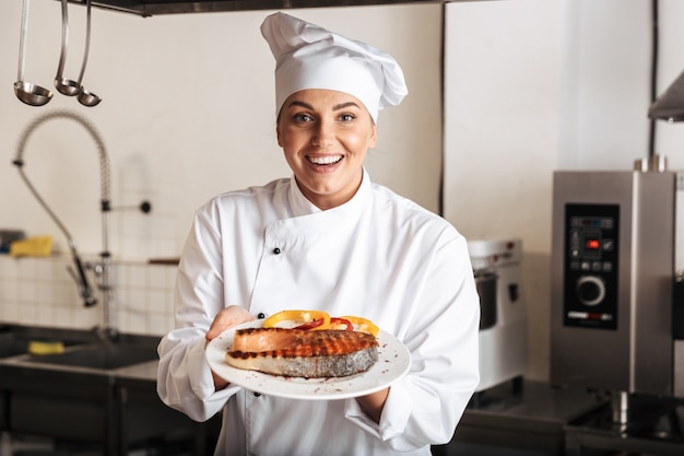 Mulher sorridente, chef cozinheira de uniforme, mostrando bife de salmão grelhado cozido em pé na cozinha