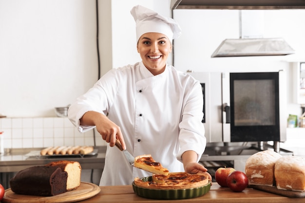 Mulher sorridente, chef cozinheira usando uniforme, mostrando torta cozida em pé na cozinha