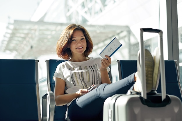 Mulher sorridente com bilhete e bagagem no aeroporto. conceito da viagem aérea com a menina ocasional nova que senta-se com a mala de bagagem de mão na porta que espera no terminal.