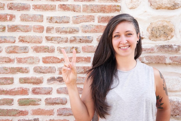 Mulher sorridente com cabelo comprido, fazendo um sinal de vitória com a mão em uma parede de tijolos