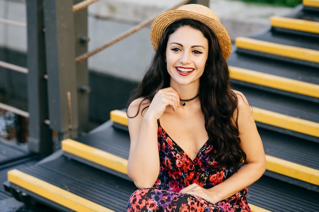 Mulher sorridente com cabelo escuro luxuoso usando vestido e chapéu de palha sentado nas escadas