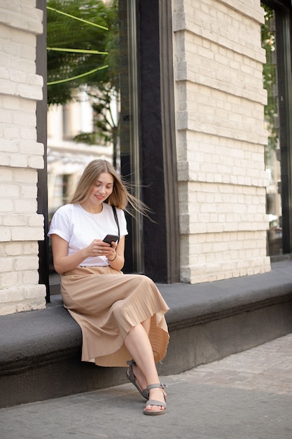Mulher sorridente com cabelo loiro em uma camiseta branca usando smartphone enquanto está sentada na rua contra o fundo de uma grande janela perto de um prédio de tijolos