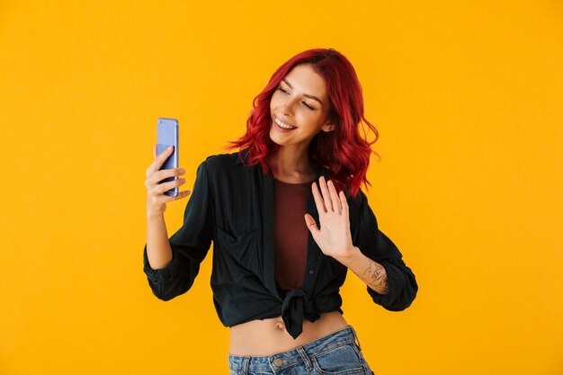 Mulher sorridente com cabelo ruivo cacheado acenando com a mão e tirando foto de selfie no celular isolado