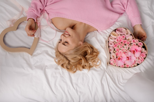 Mulher sorridente com camisa rosa, deitada na cama perto da caixa de forma de coração de flores rosas coloridas e uma capa