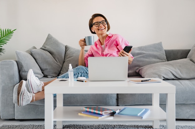 Mulher sorridente com camisa rosa relaxada bebendo chá em casa na mesa trabalhando online no laptop em casa