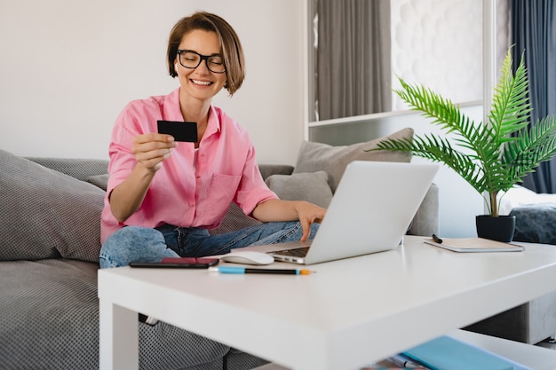 mulher sorridente com camisa rosa sentada no sofá em casa na mesa segurando carros de crédito pagando compras online no laptop de casa