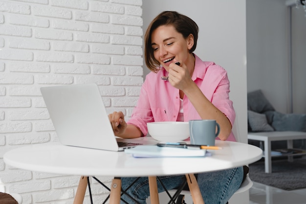 Mulher sorridente com camisa rosa tomando café da manhã em casa na mesa trabalhando online no laptop de casa, comendo cereais