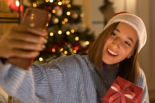 Foto mulher sorridente com chapéu de papai noel fazendo selfie enquanto segura o presente de natal