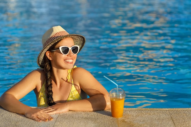 mulher sorridente com chapéu e óculos de sol, bebendo smoothies na piscina