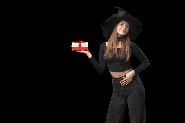 Mulher sorridente com chapéu pontudo preto segura um presente Olha para a caixa de presente da câmera Copie o espaço Menina bruxa isolada em fundo preto