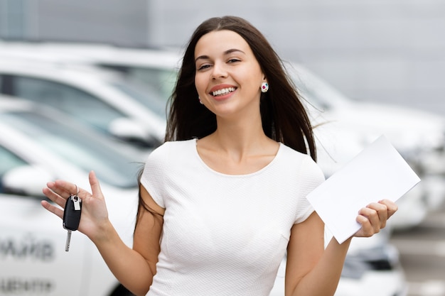 Mulher sorridente com chaves e acordo sobre plano de fundo da concessionária de automóveis