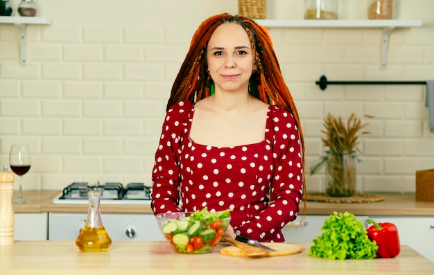 Mulher sorridente com dreadlocks no vestido com salada de legumes cozidos na cozinha Mulher positiva carinhosa olhando para a câmera em pé na mesa da cozinha