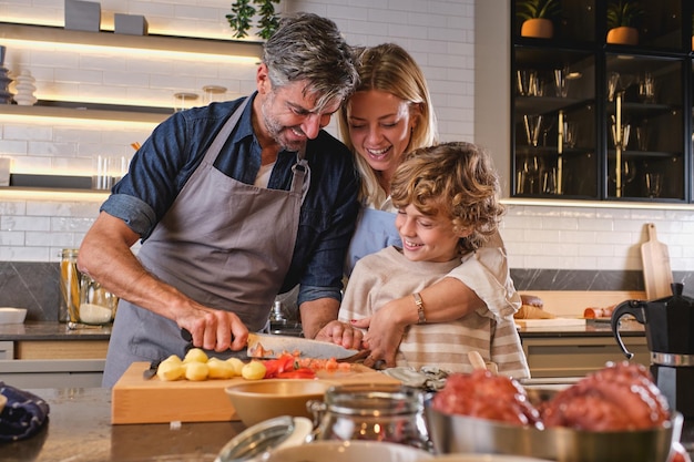 Mulher sorridente com filho bonitinho parado perto do balcão da cozinha e vendo o pai cortando tomate