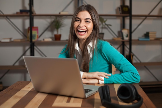 Mulher sorridente com fones de ouvido está sentada à mesa de madeira com laptop cinza em cima da mesa