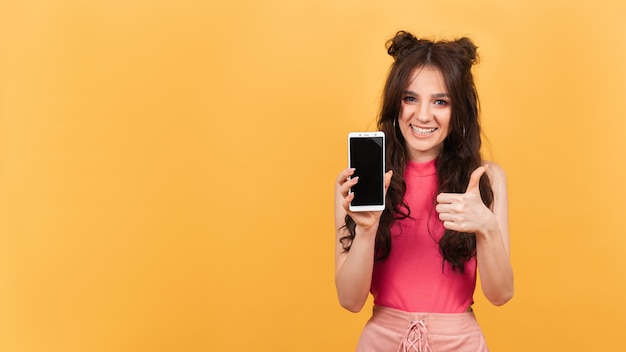 Mulher sorridente com o polegar para cima segura um telefone com tela em branco para maquete em fundo laranja