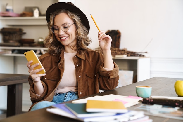 Mulher sorridente com óculos e chapéu usando o celular enquanto estudava com cadernos em casa