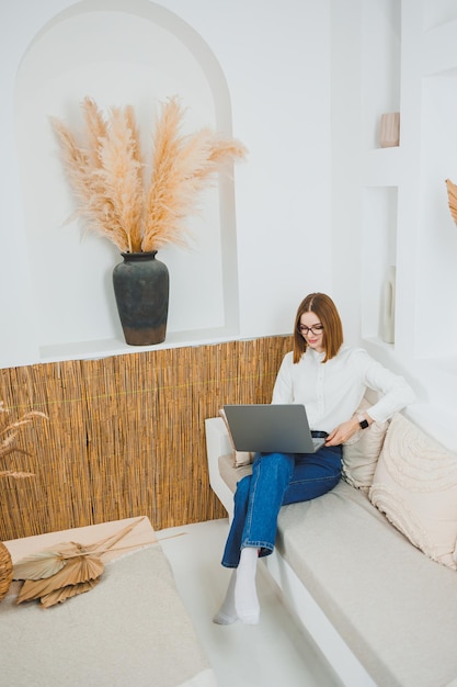 Mulher sorridente com óculos sentada no sofá em casa relaxada usando laptop trabalhando remotamente on-line vestindo camiseta branca e jeans