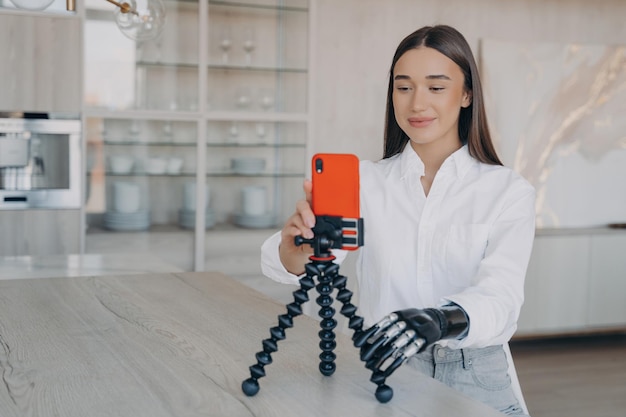 Foto mulher sorridente com um braço protético colocando um telefone celular em um tripé para uma chamada de vídeo em uma cozinha moderna