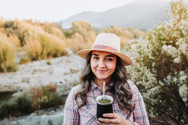 Mulher sorridente com um chapéu e uma blusa rosa pálida, bebendo mate em um espaço natural em su