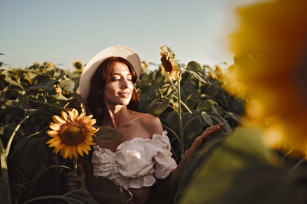 Mulher sorridente com um chapéu parada em um campo de girassóis ao pôr do sol