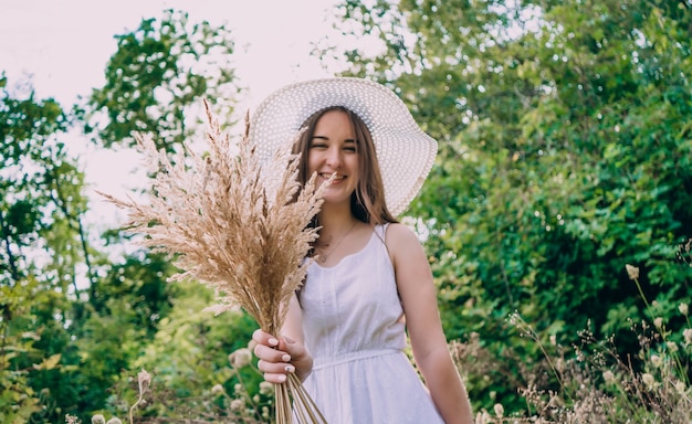 Mulher sorridente com um grande chapéu branco com um buquê em um dia ensolarado. garota feliz com um buquê de flores secas, posando em um fundo de folhas verdes.