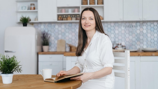 Mulher sorridente com um livro e uma xícara de café sentada em sua cozinha