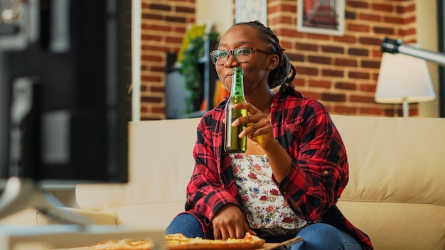 Mulher sorridente comendo uma fatia de pizza na sala de estar, curtindo um filme de comédia na televisão. Jovem se sentindo feliz bebendo garrafa de cerveja e jantando na entrega para viagem. Tiro portátil.