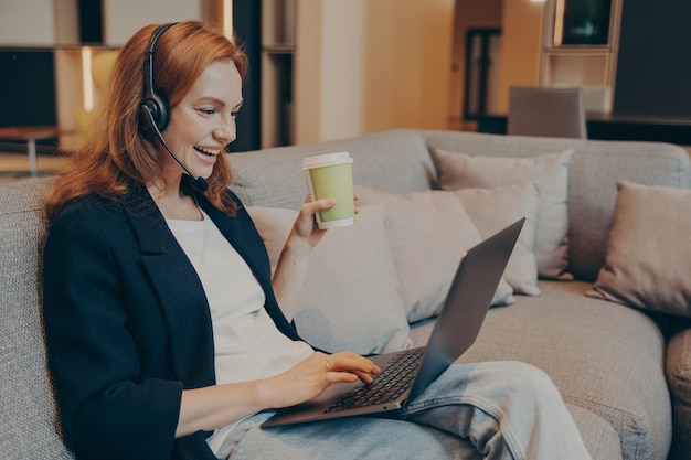 Mulher sorridente de cabelo vermelho animado com fone de ouvido sem fio e laptop sentado no sofá em um café