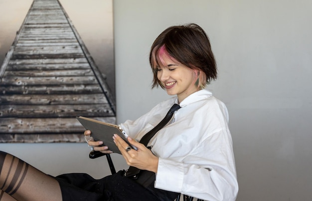 Mulher sorridente de camisa e gravata reclinada em uma cadeira, olhando para o tablet, vista horizontal lateral