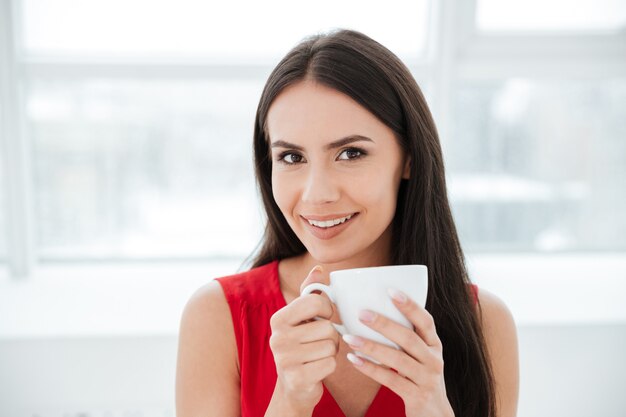 Mulher sorridente de camisa vermelha, segurando uma xícara de chá no escritório e olhando para a câmera. Janela no fundo