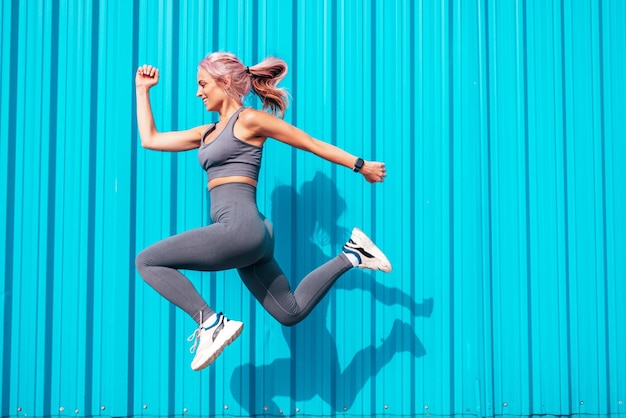Mulher sorridente de fitness em roupas esportivas cinza com cabelo rosa Jovem bela modelo com corpo perfeitoFêmea posando na rua perto da parede azulAlegre e feliz pulando e correndo