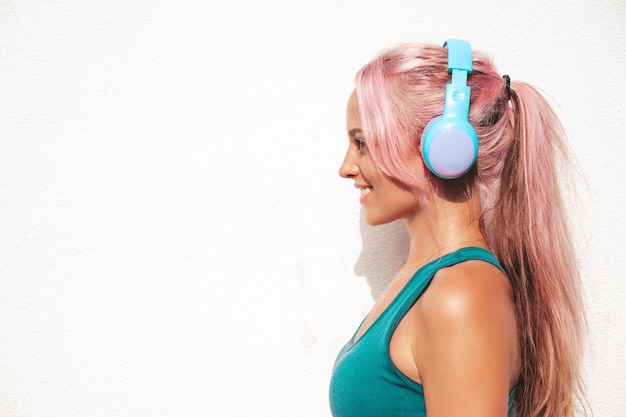 Foto mulher sorridente de fitness em roupas esportivas verdes com cabelo rosa jovem bela modelo com corpo perfeitofêmea posando na rua perto de parede branca ouvindo música em fones de ouvido sem fio