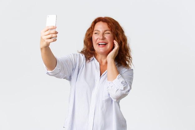 Mulher sorridente de meia-idade com cabelo vermelho tirando selfie no smartphone