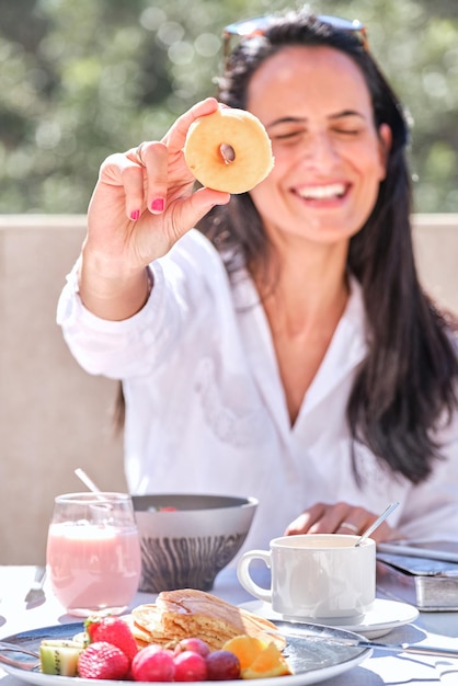 Mulher sorridente demonstrando um donut delicioso enquanto toma o café da manhã servido na mesa na varanda do café