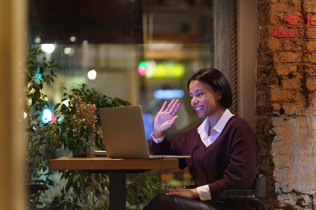 Mulher sorridente diz olá e acena com a mão para o monitor do laptop durante o trabalho de videoconferência do café