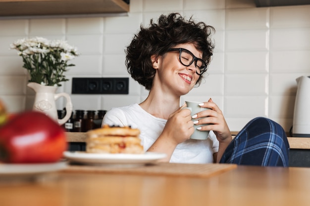 Mulher sorridente e alegre tomando um saboroso café da manhã enquanto está sentada na cozinha de casa, bebendo café